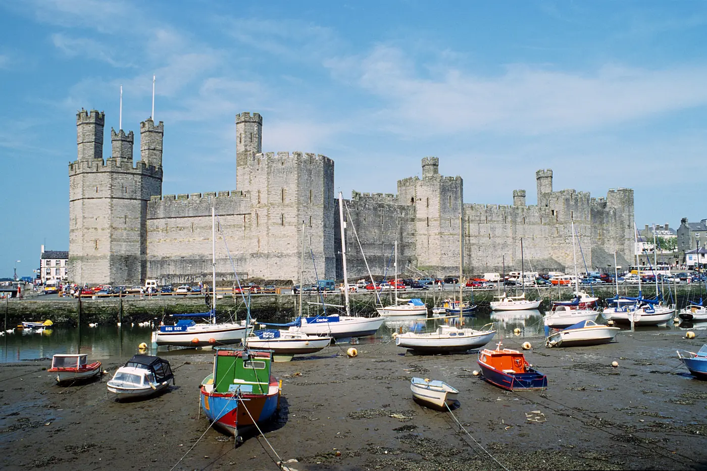 Caernarfon Castle seen across the harbour at low tide, its polygonal towers and banded stone walls above moored boats